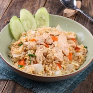 A bowl of chicken fried rice with fresh vegetables on a rustic wooden table.